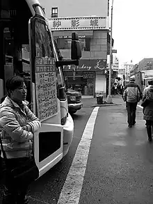 The black-and-white image shows. To the left of the image, a woman of East Asian extraction standing next to a coach with a sign on its door advertising prices of journeys from New York to Washington D. C. in Han characters. On the right, pedestrians walk across the road in front of the coach under the guidance of a green traffic light. In the left background on the street corner is a Chinese bakery. In the right background there is visible a crowded sidewalk, alongside which three white delivery trucks are parked, and a street of square high-rise apartment buildings.