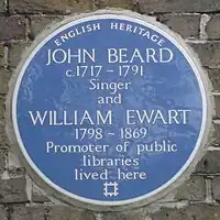 A Blue plaque on a brick wall with the words "John Beard C1717 – 1791 Singer and William Ewart 1798 – 1861 Promoter of Public Libraries