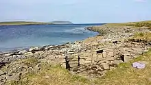The remains of a Pictish building made of stone. The ruins stand near the landward edge of a stony beach, where the ground transitions from stones to grass. Water is visible beyond the beach, and there is a land visible on the horizon.