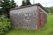 A small wooden building, with a slanting shed roof. One window is visible on one side, a Dutch door on the other, with a horseshoe nailed to the door. The building has red wooden trim around the corners, window and door, and the exterior walls are covered with grey wood shingles.