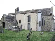A small stone church seen from the south-east with a porch to the left, a protruding rendered south aisle in the middle, and a short chancel to the right. On the west gable is a bellcote, and in front of the church is a churchyard cross