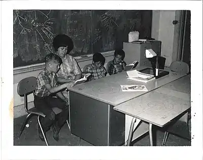 Three young boys sit at a table with their teacher. The teacher points to the words that one boy is reading in his textbook.