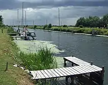 A broad, flat artificial cut with dark water and bright green raised banks.  A patch of green algae stains the water.  Several modern yachts are moored on wooden jetties spaced along the bank