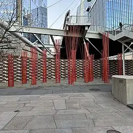 An outdoor urban installation consisting of multiple thin red rods arranged in clusters, creating a striking visual contrast against the gray paving stones, a brick wall, and the metal framework of an overhead pedestrian bridge.