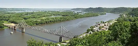 Summertime overlooking a wide and winding river. A small town is to the right. An old steel cantilever-truss bridge dominates the foreground.