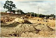 Photograph of construction of the Westgate Shopping Centre in Massey, Auckland, New Zealand, in 1994. The image depicts a pit of dirt with construction vehicles, including several trucks and a bulldozer.