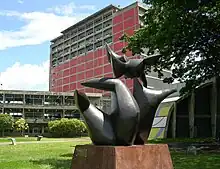 Escultura ao ar livre moderna, com um edifício vermelho e passarela elevada ao fundo