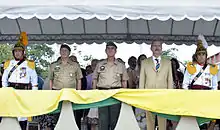 Aldo aparece em posição de sentido num estande, de terno, junto de dois outros comandantes militares e cercados por dois soldados em uniforme tradicional