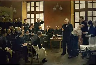 Uma pintura de uma palestra médica do século XIX. Na frente da classe, uma mulher desmaia nos braços de um homem parado atrás dela, enquanto outra mulher, aparentemente uma enfermeira, estende a mão para ajudar. Um homem mais velho, o professor, está ao lado dela e gesticula como se estivesse fazendo uma observação. Duas dúzias de estudantes do sexo masculino os observam.