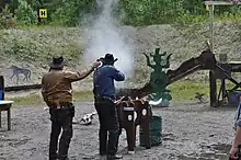 Um atirador de CAS disparando um rifle de alavanca em alvos de aço. O Range Officer à esquerda está segurando um cronômetro para medir o tempo.
