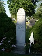 Foto de um túmulo com dois vasos de flores e uma bandeira branca ao seu lado. Ao seu redor, há árvores e outros túmulos. Em seu epitáfio, traduzindo de inglês para português, está escrito: Em memória. Sojourner Truth. Nasceu escravizada no Condado de Ulster, Nova Iorque no século 18. Morreu em Battle Creek, Michigan, em 26 de novembro de 1883. Com idade aproximada de 105 anos. "Deus está morto".