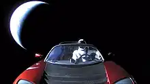Photograph of the front of a red convertible sports car floating in space.  There is a humanoid figure in the driving seat.  In the background, partially illuminated in a crescent shape, is planet Earth.
