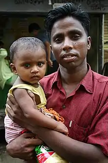 A young father dressed in a pink cotton shirt holds his child and gazes at the câmera looking proud but tired. The little girl, wearing a sleeveless dress, sits on her father's arm and frowns directly at the câmera.