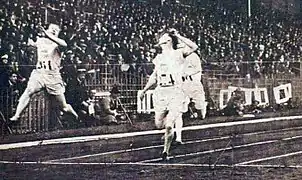 Foto de vários homens perto de cruzar a linha de chegada de uma corrida, um está no ar depois de um salto.