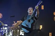 Uma imagem de um homem vestindo uma camisa preta e calça está tocando enquanto segura uma guitarra preta