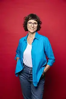 Imagem de corpo inteiro feita em estúdio. Heloisa sorri. Usa óculo, blusa azul, camiseta branca e calça jeans,