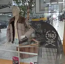 Foto colorida de uma vitrine, dentro com um boneco manequim vestindo uma jaqueta de pele ao lado de um leme de cauda de avião. O leme apresenta várias marcações, no topo o número 150, rodeado por uma coroa de flores. Abaixo da coroa há duas espadas cruzadas e numerosas listras brancas.
