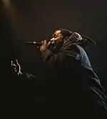 A black man, dressed in all black, stands side on to the camera. He is performing on stage with a microphone in his hand