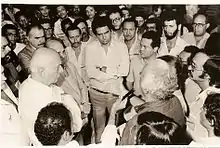 Fotografia em preto e branco, mostra Tancredo Neves, o candidato civil às Diretas, em 1984, recendo documento entregue por trabalhadores, com a presença de muitas personalidades.