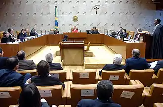 Em primeiro plano, as cadeiras de espectadores do julgamento, todos trajados formalmente. Ao fundo e centralizada, a presidente do STF Ellen Gracie; à direita dela, o PGR e a sua esquerda um assessor judiciário. Atrás dela, a bandeira do Brasil, o emblema nacional e um crucifixo cristão. Os outros ministros se dispõem lateralmente. A direita, o ministro Barbosa está de pé, em frente de um púlpito com microfone, lendo.