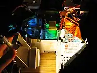 Acima um conjunto ótico de um projetor aberto, mostrando o funcionamento dele. Foi colocado uma lâmpada comum para exemplificar como a luz é transmitida dentro do projetor, passando pelas lentes, espelhos e filtros dicróicos.