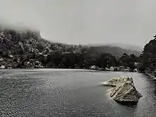 El agua contiene decenas de ondas provocadas por el viento. Al fondo de la foto se ve la cima de la montaña tapada por la niebla, que desciende hasta casi tocar el agua. La ladera de la montaña está manchada por trozos de nieve.