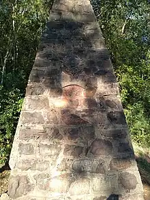 Obelisco de pedras basálticas acinzentadas contendo o brasão de armas do Brasil e a inscrição 1902.