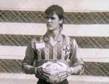 Um goleiro jovem, segurando uma bola de futebol com as duas mãos, olhando para longe, com cara de concentrado