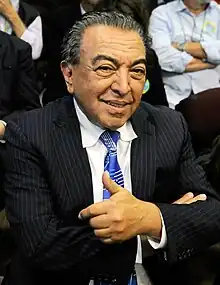 Foto de Maurício de Sousa. Ele é um homem idoso, com cabelo ainda preto entre cabelos brancos, e retraído para trás. Ele está vestido com um terno preto de listas verticais cinzas, uma blusa social branca e uma gravata azul com detalhes cor-de-prata. Ele sorri, enquanto levanta o polegar em um sinal de joia.