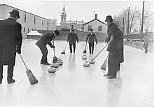 Um grupo de homens, vestidos com roupas de inverno e usando chapéus, participam de uma partida de curling. Um deles varre o gelo, enquanto os outros, também com vassouras na mão, observam.