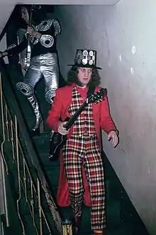 A man walks down a flight of stairs. He holds a guitar and wears a black hat studded with round metal mirrors. His waistcoat and trousers are tartan. His coat and shirt are plain red. His socks are striped.