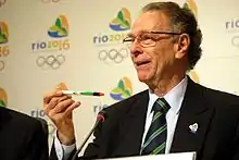 Imagem que mostra Carlos Arthur Nuzman sorrindo à frente de um microfone. No fundo, há o símbolo das Olimpíadas do Rio de Janeiro de 2016.