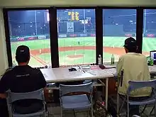 Cabine do anotador oficial, com duas pessoas observando uma partida de beisebol