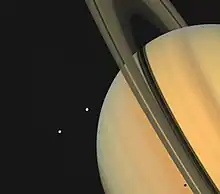 Saturno no canto direito da imagem, coloração bege com faixas marrons claro. Anéis marrons inclinados, projetam sombra escura no planeta. Próximo do planeta, à esquerda, dois pontos brancos.