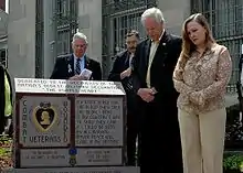 Uma foto colorida de Daniel e Maureen Murphy ao lado de um monumento em frente à Agência Postal dos Estados Unidos Tenente Michael P. Murphy em Patchogue, Nova Iorque. O monumento tem uma Medalha Purple Heart e algumas palavras gravadas nele e há dois homens ao fundo.