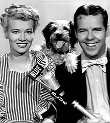 Penny Singleton as Blondie and Arthur Lake as Dagwood Bumstead, from a 1944 publicity photograph. A smiling white woman with blonde curls and a striped pinafore apron; a smiling white man wearing a bow tie; a small dog posed between their shoulders; and a microphone in front of them, labeled "Blue".