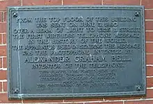 An image of darkened brass historical plaque with a streak of green corrosion running down it, mounted on the exterior side of a brick building.