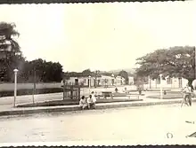 A Praça Coronel Collaço, década de 1950. Sentados, Irma Gerlach Collaço e seu marido, o farmacêutico Newton de Andrade Collaço, com a filha Maria Christina. Ao fundo identifica-se o Armazém Soares, de Satiro Soares. A figueira já está com vistoso porte