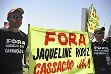 Dois homens de preto seguram uma grande faixa amarela onde se lê "FORA Jaqueline Roriz Cassação Já!"