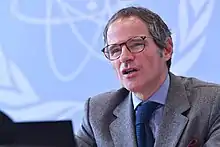 A man in glasses speaks while sitting in front of a blue background