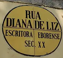 Placa oval cerâmica de fundo amarelo com linhas azuis, onde está inscrito o texto "Rua Diana de Liz. Escritora Eborense. Sec XX".