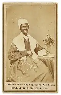 Foto de um carte-de-visite de uma mulher por volta dos 60 anos, em um vestido longo, tricotando um cachecol ao lado de uma mesa com um vaso de flor. Na parte de baixo da carte, traduzindo de inglês para português, está escrito: Eu vendo a sombra para sustentar a substância. Sojourner Truth.