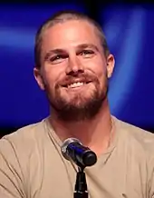 Uma fotografia de Stephen Amell falando em uma convenção atrás de um microfone