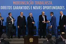 No topo da imagem, está escrito em letras maiúsculas num fundo azul "modernização trabalhista". Abaixo dela, está escrito "direitos garantidos e novas oportunidades". Encontra-se da esquerda pra direita, Ronaldo Nogueira, Eliseu Padilha, Michel Temer, Henrique Meirelles e Romero Jucá.