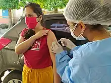 Foto de Carol Cardoso tomando uma vacina. Ela está vestida de máscara e blusa vermelha, com calças amarelas. Atrás dela, há um carro de cor metálica. Ao lado de Carol, a enfermeira que está aplicando a injeção está com uma touca para cabelo, máscara e luvas brancas, e veste-se de azul bebê. Carol é parda, de olhos e cabelos pretos, e seu cabelo é liso e longo.