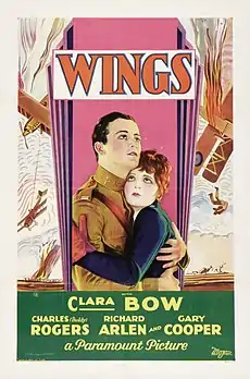 Cartaz promocional de um filme. Ele se concentra em duas pessoas. O homem está vestindo um terno de aviador e a mulher está vestindo uma jaqueta azul e luvas vermelhas.