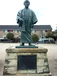 Photo couleur d'une statue de bronze sur un socle en pierre figurant un homme debout en habits traditionnels japonais.